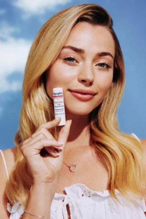 Smiling woman with long blond hair holds Aquaphor lip balm tube against a blue sky background. Close-up beauty portrait shot.