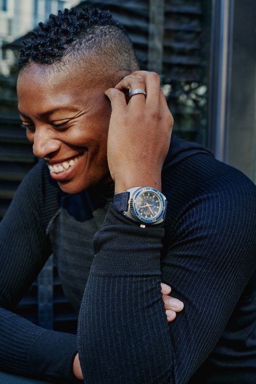 Close-up portrait of a Black man with short dark hair wearing a ribbed black top and a blue wristwatch, smiling while touching his head.