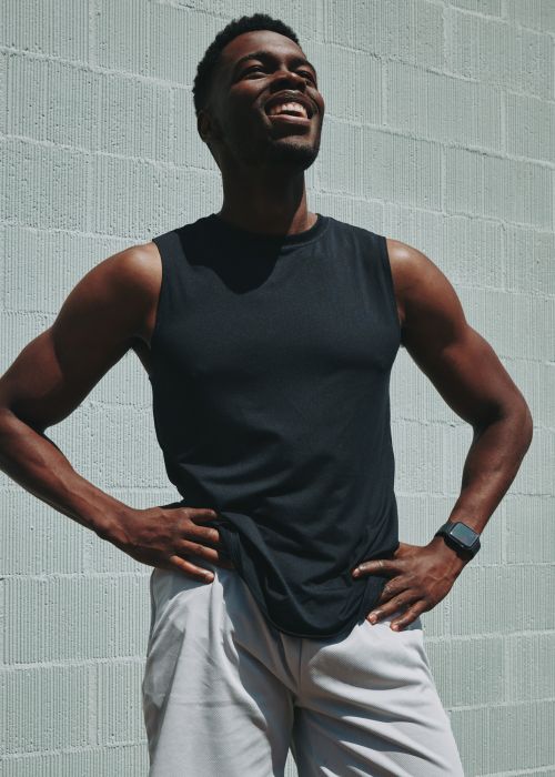 A smiling Black man in a sleeveless shirt poses outdoors against a textured white wall, showcasing fitness and casual style.