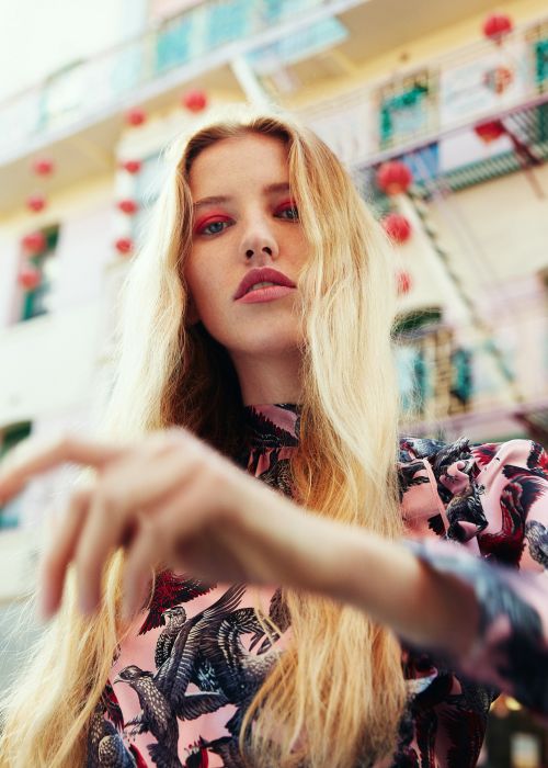 Young woman with long blonde hair poses in a patterned dress in front of a cityscape with hanging red lanterns, beauty style.