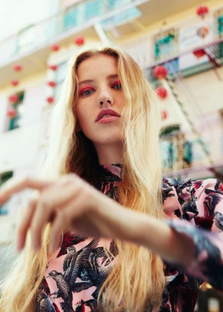 Young woman with long blonde hair poses in a patterned dress in front of a cityscape with hanging red lanterns, beauty style.