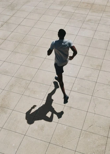 An adult male is jogging outdoors on a tiled surface, captured from an overhead angle casting a distinct shadow next to him.