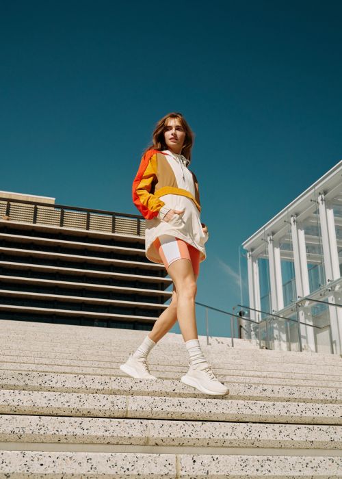 Woman in color-block jacket walks down stairs on a sunny day near modern building with blue sky in an eye-level, medium shot.