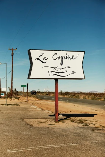 Vintage 'La Copine' sign in the desert under a clear blue sky. An empty asphalt parking lot in the foreground.