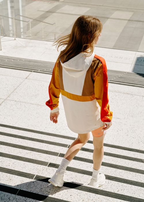 A woman walks down a set of outdoor stairs, wearing a color-blocked anorak, peach shorts, white athletic shoes, and white socks in sunlight.