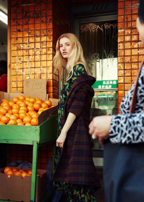 A blonde female model in a printed dress and plaid coat stands next to oranges on a city street with passersby and orange brick walls.