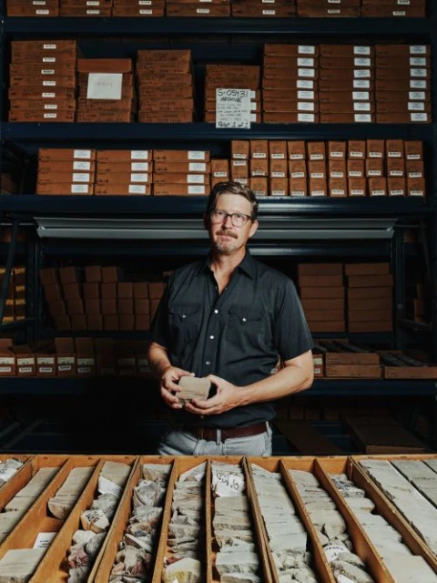 Man in glasses holds rock core sample in front of rock core samples in a drawer and stacked boxes on shelving in an indoor setting.