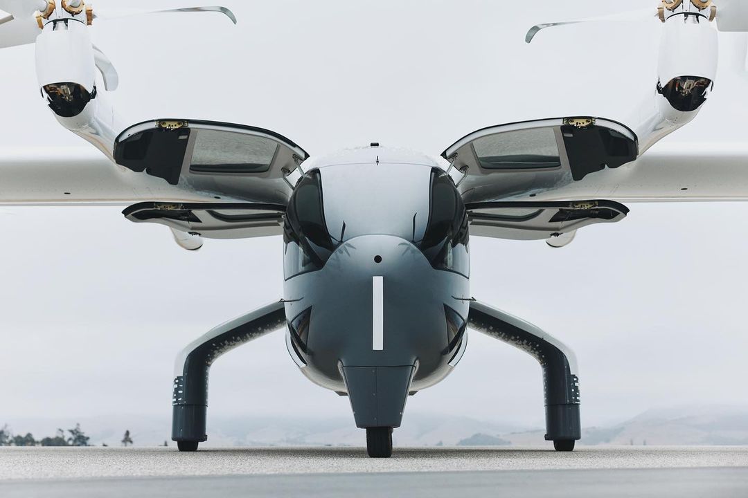 Front view of an electric vertical takeoff and landing (eVTOL) aircraft with rotors, landing gear, and sleek gray and white design.