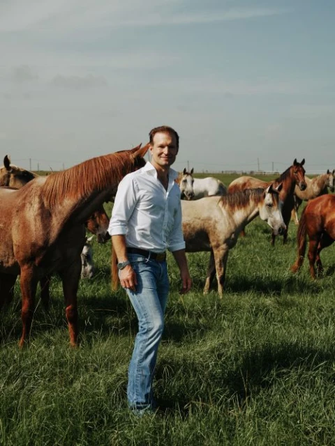 A man in a white shirt and jeans stands in a grassy field surrounded by a herd of horses under a bright, cloudy sky. 