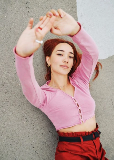 Woman lying on concrete with red hair, pink top and red pants, arms above her head, looking at camera. Portrait orientation.