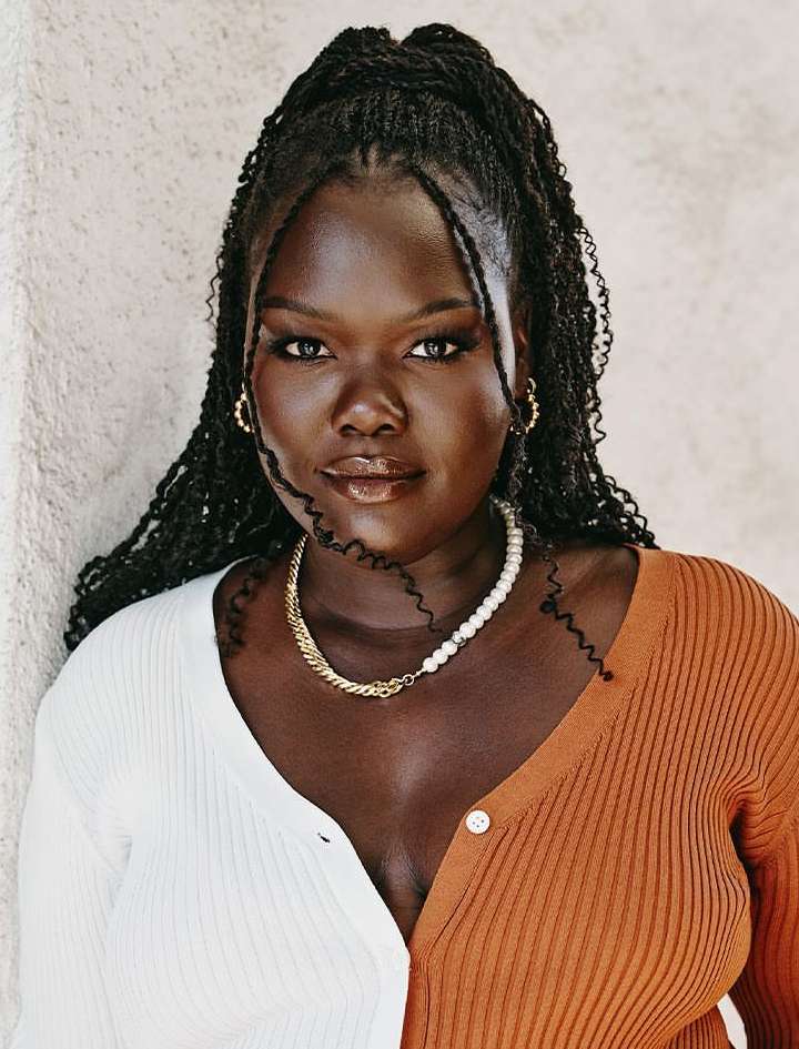 Portrait of a smiling adult female with braided hair wearing a two-tone cardigan with pearl and gold necklaces against a cream backdrop.