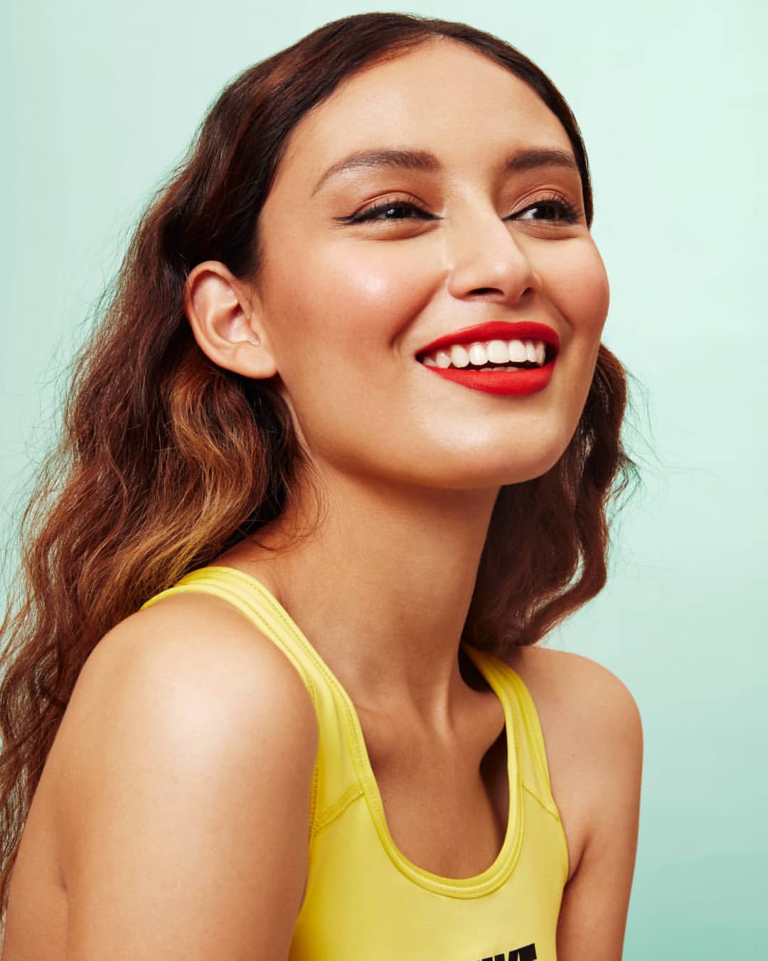 Portrait of a brunette woman in a yellow tank top with red lipstick smiling against a light teal background in a studio setting.