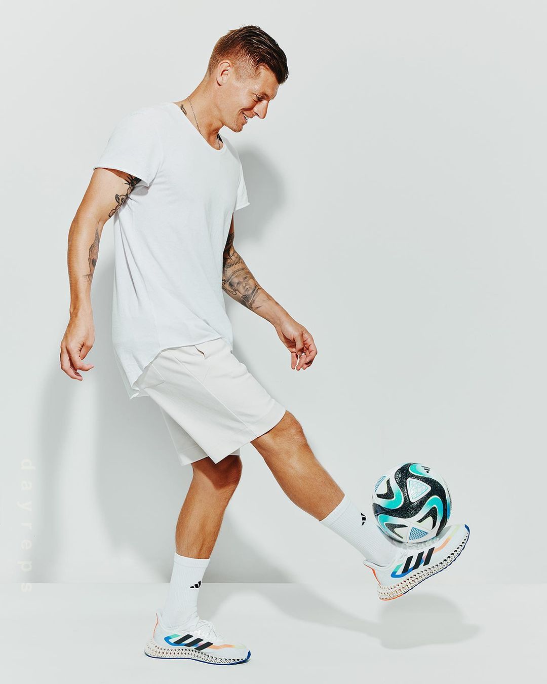 Full shot of an adult male playing with a soccer ball in a white t-shirt and shorts in a brightly lit studio environment.