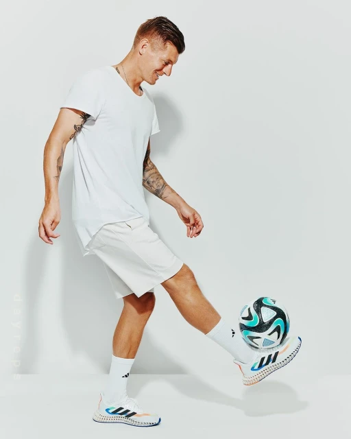 Full shot of an adult male playing with a soccer ball in a white t-shirt and shorts in a brightly lit studio environment.