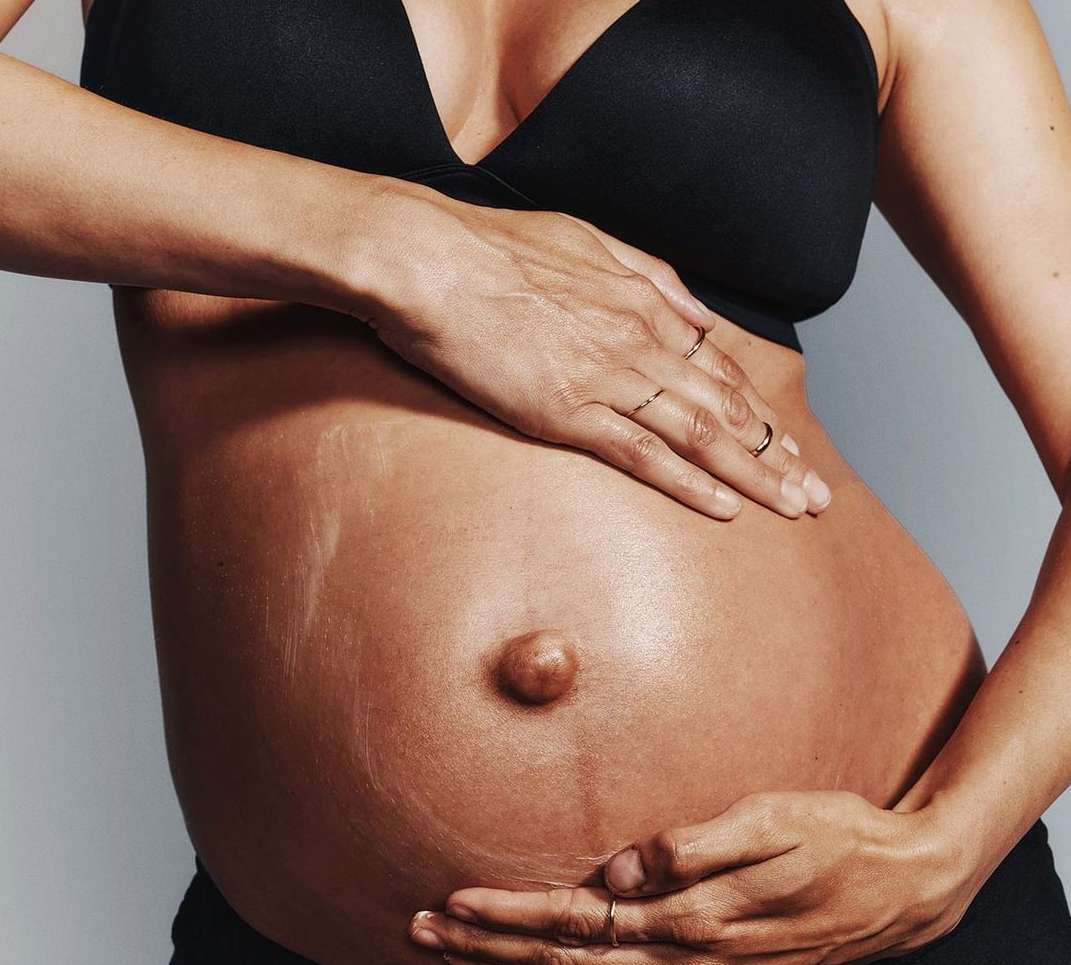 Close-up studio shot of pregnant woman’s bare belly with hands, black bra, and seamless gray background.