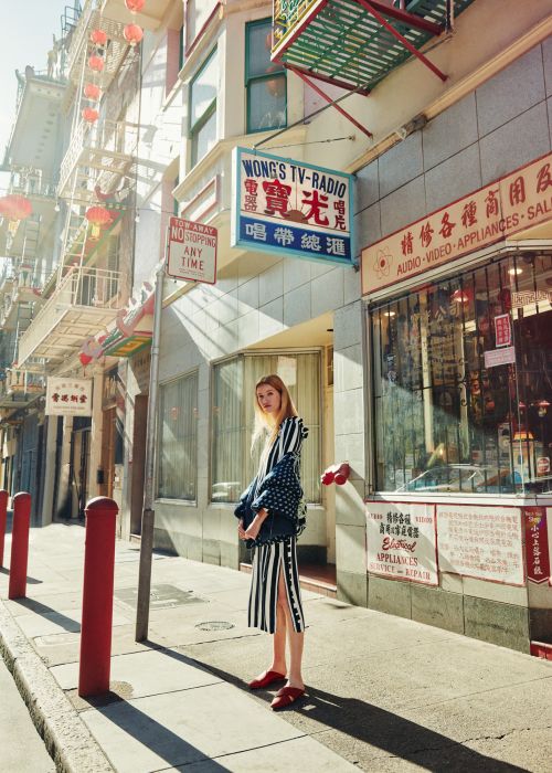 Fashionable adult woman stands on a city sidewalk in front of shops and buildings on a sunny day. She wears a jacket and striped dress.