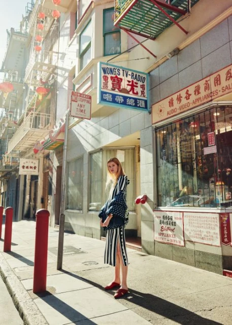 Fashionable adult woman stands on a city sidewalk in front of shops and buildings on a sunny day. She wears a jacket and striped dress.