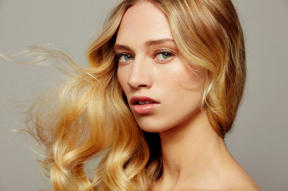 Close-up portrait of a woman with long blonde wavy hair blowing in the wind against a solid grey background