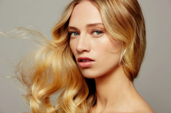 Close-up portrait of a woman with long blonde wavy hair blowing in the wind against a solid grey background