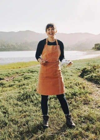 Woman in orange apron standing on grassy shore with lake and hills in background
