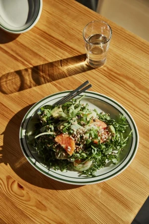 Close-up of a vibrant salad with citrus, greens, and seeds served on a plate with a glass of water on a wooden table.