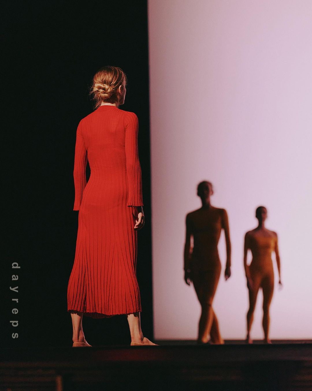 A woman in a red dress faces away from the camera, with two blurred figures in the background on a stage bathed in bright light.