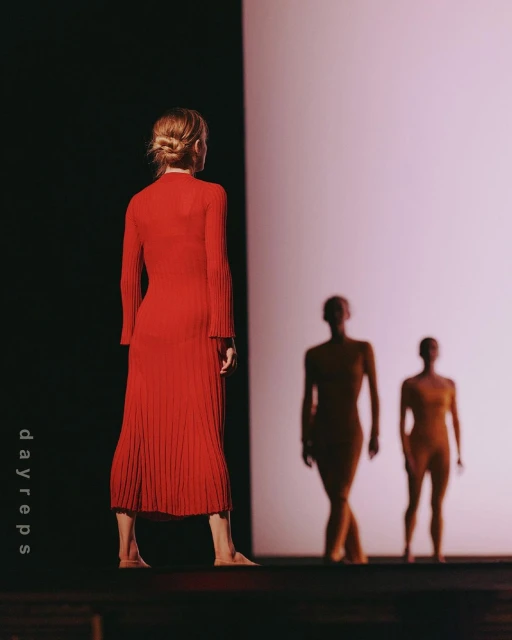 A woman in a red dress faces away from the camera, with two blurred figures in the background on a stage bathed in bright light.