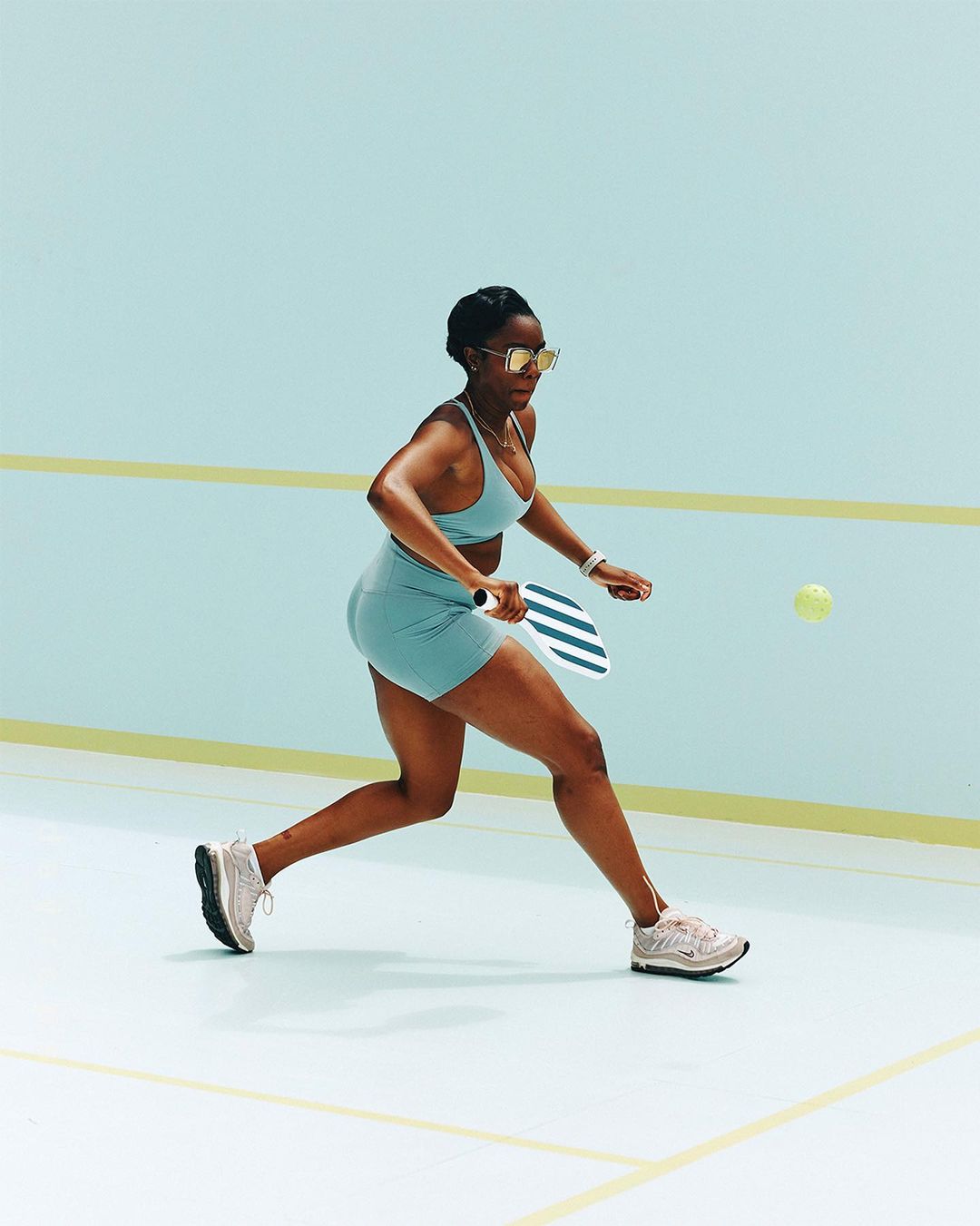 African American woman in sportswear plays pickleball indoors. The athlete wears sunglasses and aims to hit a yellow ball with a paddle.