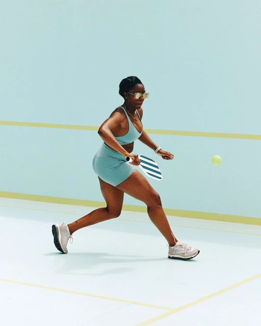 African American woman in sportswear plays pickleball indoors. The athlete wears sunglasses and aims to hit a yellow ball with a paddle.