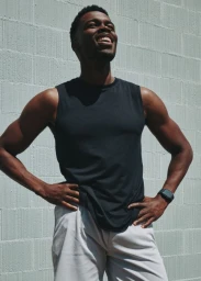 Black man in a black tank top and white shorts laughing outdoors against a light grey block wall