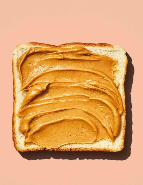 Close-up studio shot of a slice of white bread covered with creamy peanut butter on a pink background, food photography.