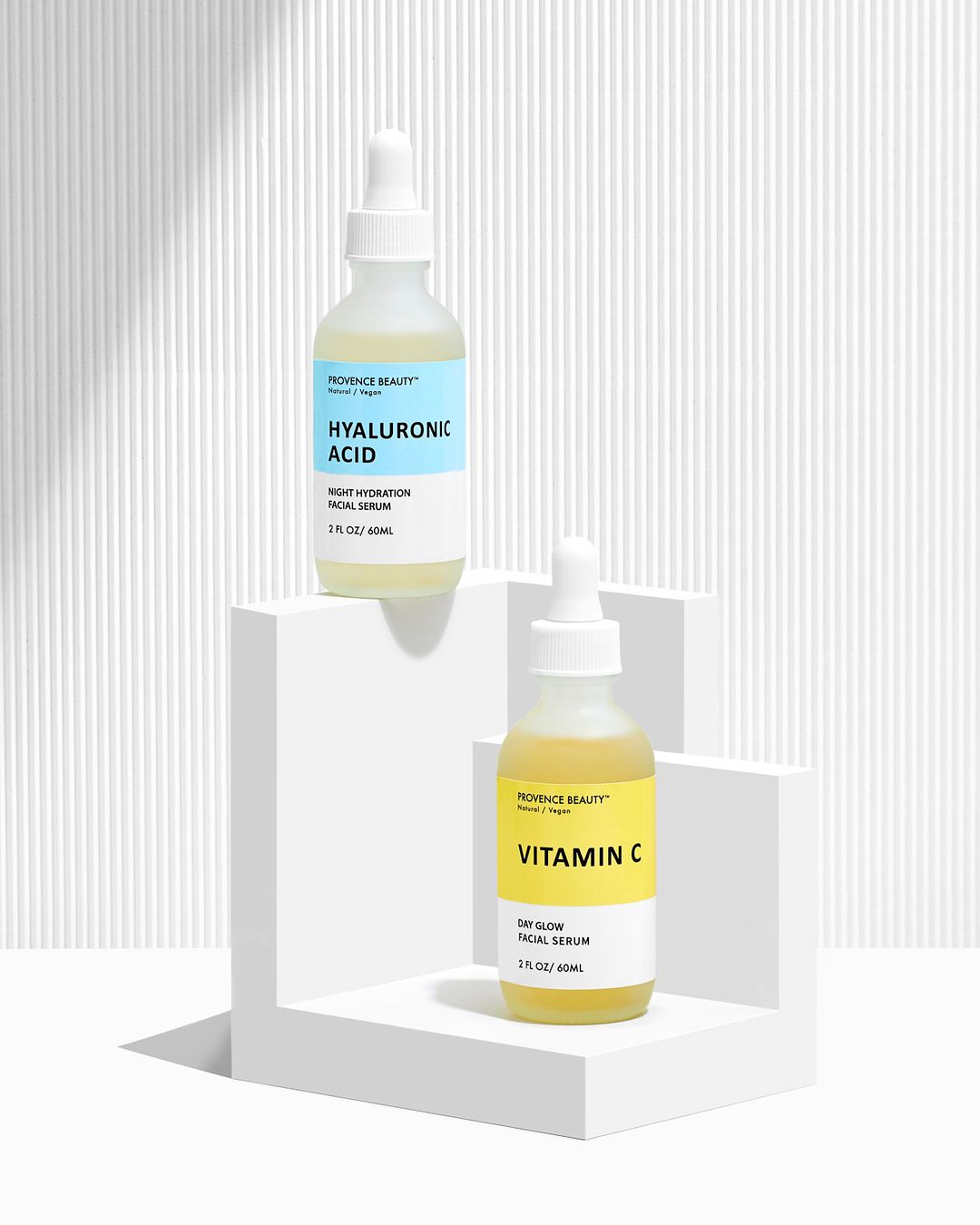 Two Provence Beauty serum bottles, one with hyaluronic acid and the other with vitamin C, are displayed on a white L-shaped platform.