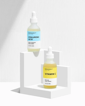 Two Provence Beauty serum bottles, one with hyaluronic acid and the other with vitamin C, are displayed on a white L-shaped platform.