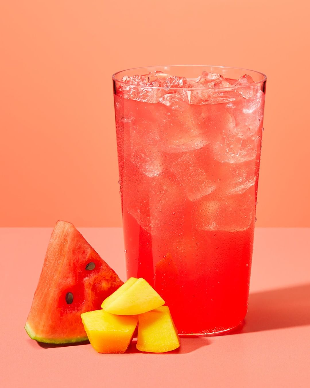 A glass of pink watermelon juice with ice sits next to a watermelon slice and chunks of mango against a peach-colored background.