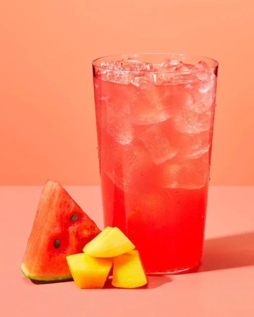 A glass of pink watermelon juice with ice sits next to a watermelon slice and chunks of mango against a peach-colored background.