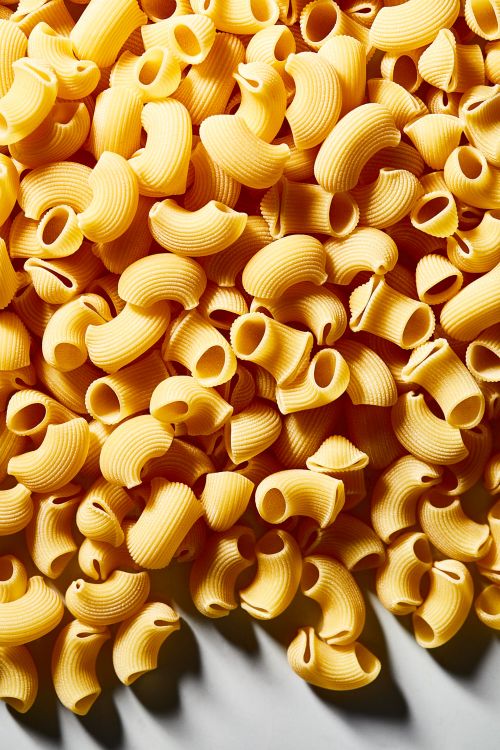 Close-up of elbow macaroni pasta showing its curved shape, ridges, and golden-yellow color. The pasta is piled randomly on a surface.