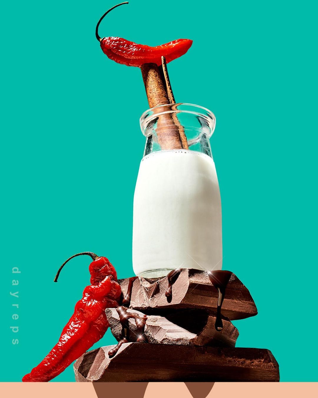 Milk in a glass bottle sits atop dark chocolate, garnished with chili peppers, against a vibrant turquoise background in a studio setting.