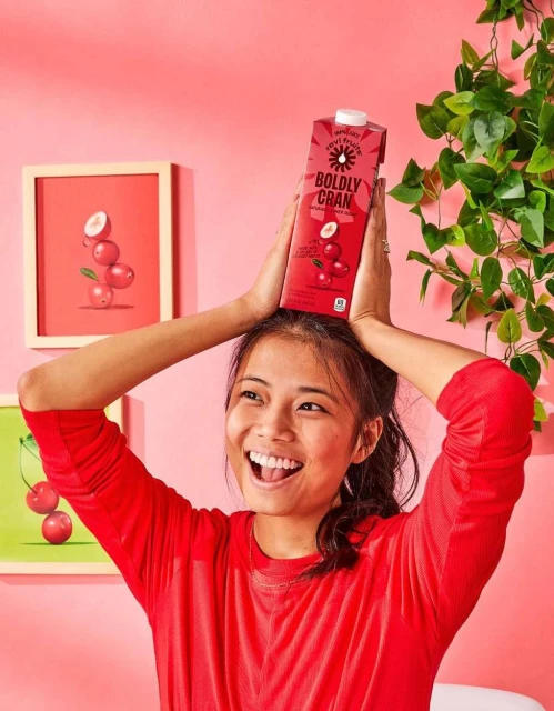 Smiling woman in red holds a Boldly Cran cranberry juice carton on her head indoors with plant and fruit artwork in background.
