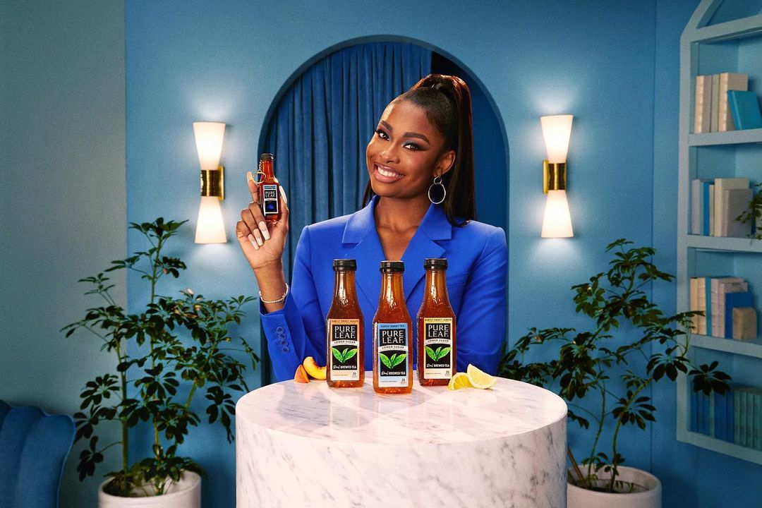 Attractive woman in a blue suit smiles, holding a drink bottle, with Pure Leaf iced tea bottles displayed on a marble table.
