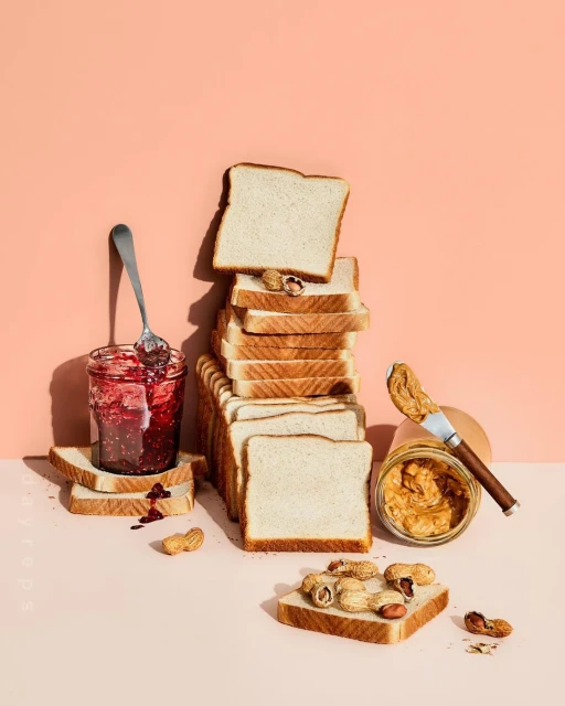 Still life featuring stacked bread slices, jars of peanut butter and jelly, and scattered peanuts on a soft pink surface.