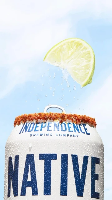 A close-up shot of a can of Native beer with a lime wedge, condensation, and a chili-salt rim, set against a cloudy blue sky.