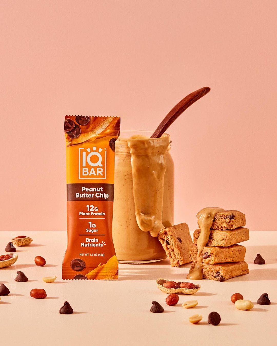 Product shot of an IQBAR Peanut Butter Chip bar, pieces of the bar, a jar of peanut butter with a spoon, peanuts, and chocolate chips.