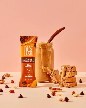 Product shot of an IQBAR Peanut Butter Chip bar, pieces of the bar, a jar of peanut butter with a spoon, peanuts, and chocolate chips.