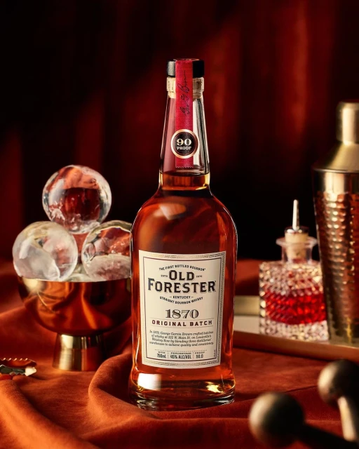 A bottle of Old Forester bourbon whiskey sits on a textured rust-colored surface with ice and cocktail equipment in the background.