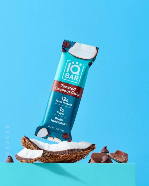 A teal-colored IQ Bar with toasted coconut chip flavor floats above a chunk of coconut and chocolate chips against a solid blue backdrop.