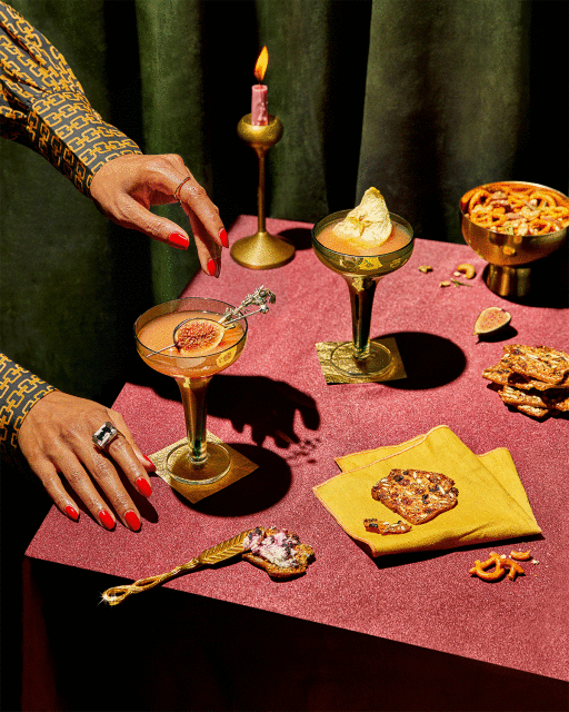 Elegant cocktail setup on a table featuring golden-stemmed glasses, figs, pretzels, and decorative spoons, under warm lighting against a dark green backdrop.