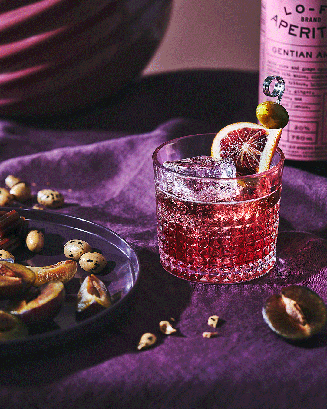 Close-up shot of a cocktail with ice and a slice of orange, accompanied by a plate of plums and nuts, set against a purple backdrop.