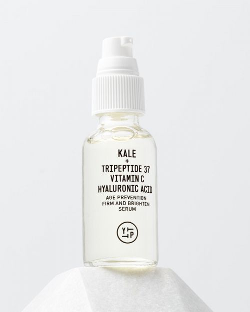 Clear glass bottle with white pump containing kale, tripeptide, vitamin C, and hyaluronic acid on white geometric pedestal.