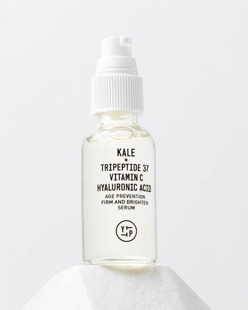 Clear glass bottle with white pump containing kale, tripeptide, vitamin C, and hyaluronic acid on white geometric pedestal.