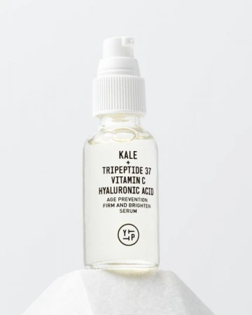 Clear glass bottle with white pump containing kale, tripeptide, vitamin C, and hyaluronic acid on white geometric pedestal.